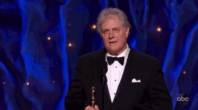 a man in a tuxedo and bow tie holds an oscar trophy in front of an abc logo