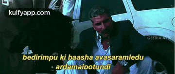 a man in a suit is sitting in front of a car and talking to someone in a foreign language .