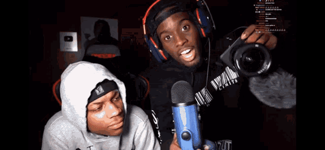 a man wearing headphones holds a blue microphone while another man looks on