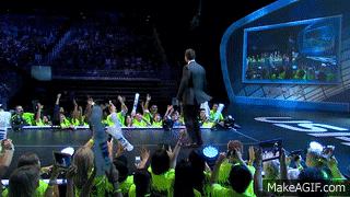a man in a suit stands on a stage in front of a crowd with a makeagif.com watermark