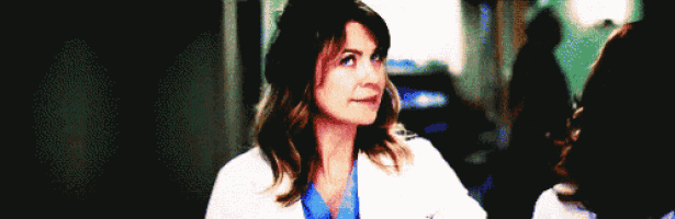 a woman in a lab coat and scrubs is standing in a hallway looking at the camera .