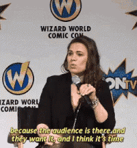 a woman speaking into a microphone in front of a wizard world comic con sign
