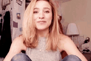 a woman in a grey tank top is sitting on a bed with her legs crossed and smiling .