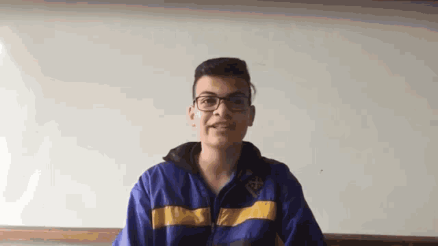 a young man wearing glasses and a blue and yellow jacket is standing in front of a whiteboard .