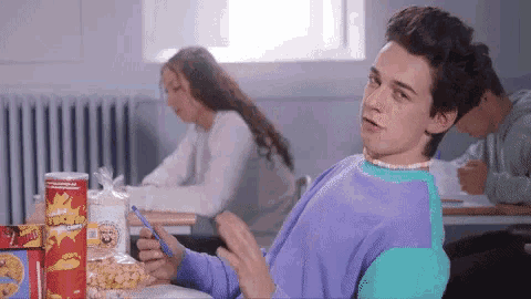 a boy in a purple sweater is sitting at a desk in a classroom with a bag of cheetos in front of him
