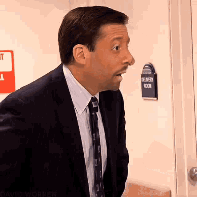 a man in a suit and tie stands in front of a delivery room sign .
