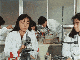 a woman is looking through a microscope in a lab while a group of people work in the background .