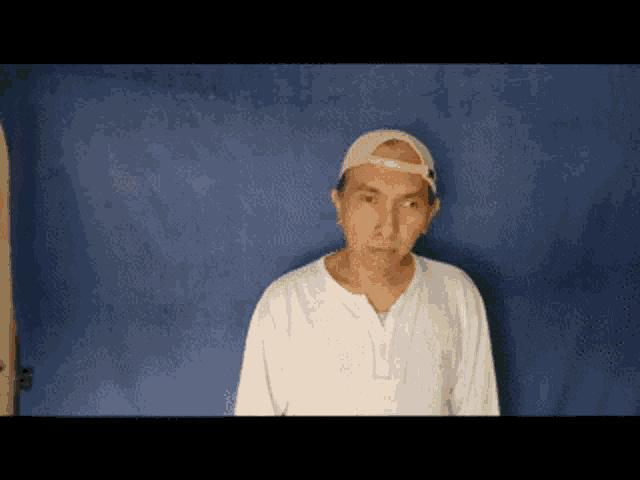 a man wearing a baseball cap and a white shirt stands in front of a blue wall