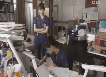 a woman in scrubs is standing next to a man sitting at a desk in a messy office .