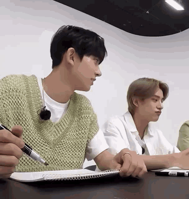 a man is sitting at a table writing in a notebook while another man looks on .