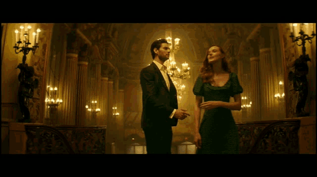 a man in a suit and a woman in a green dress are standing in a room with a chandelier