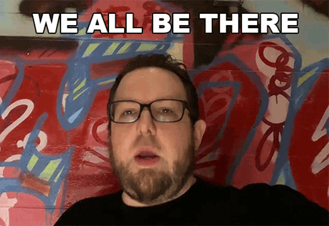 a man with glasses and a beard says we all be there in front of a graffiti wall