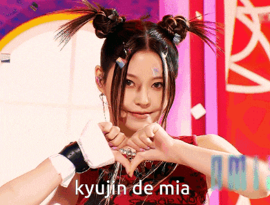 a woman making a heart shape with her hands with kyujin de mia written on the bottom right