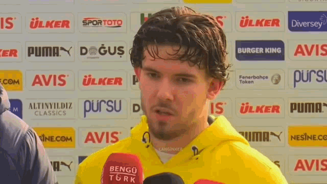 a man wearing a yellow hoodie is talking into a microphone in front of a wall of advertisements including burger king