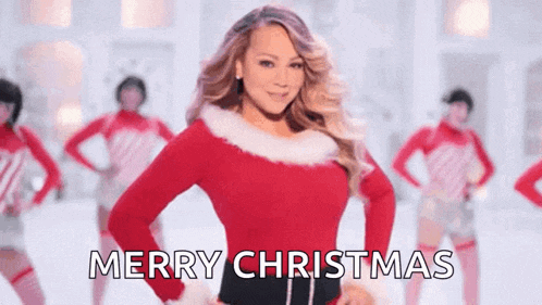 a woman in a santa claus costume is standing in front of a group of dancers and saying merry christmas .