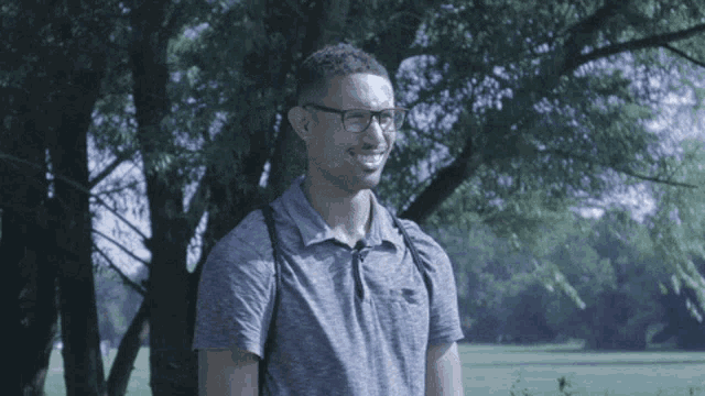 a man wearing glasses and a grey shirt is smiling in front of a tree