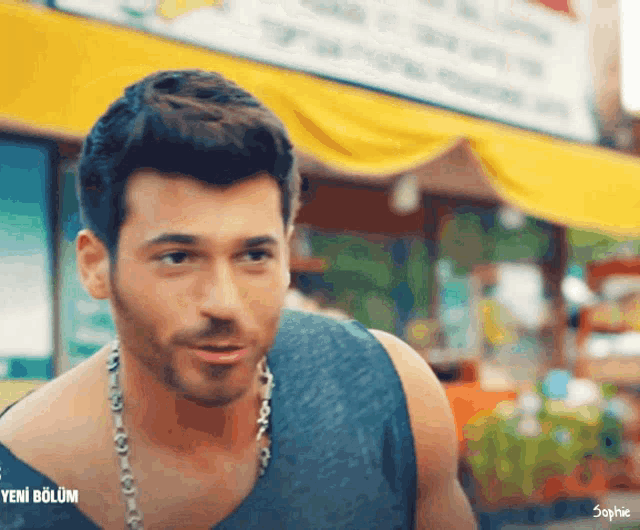 a man wearing a tank top and a necklace is standing in front of a yellow banner that says yeni bolum