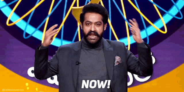 a man in a suit and turtleneck is standing in front of a sign that says now .