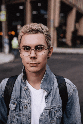 a young man wearing glasses and a denim jacket stands on a street