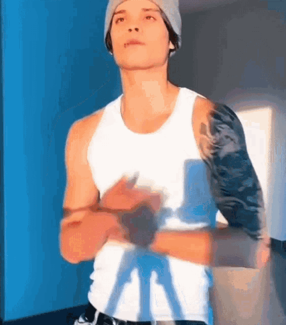 a man with a tattoo on his arm is wearing a white tank top and a gray hat .