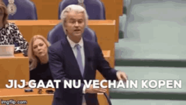 a man in a suit and tie is giving a speech in a parliament while a woman sits behind him