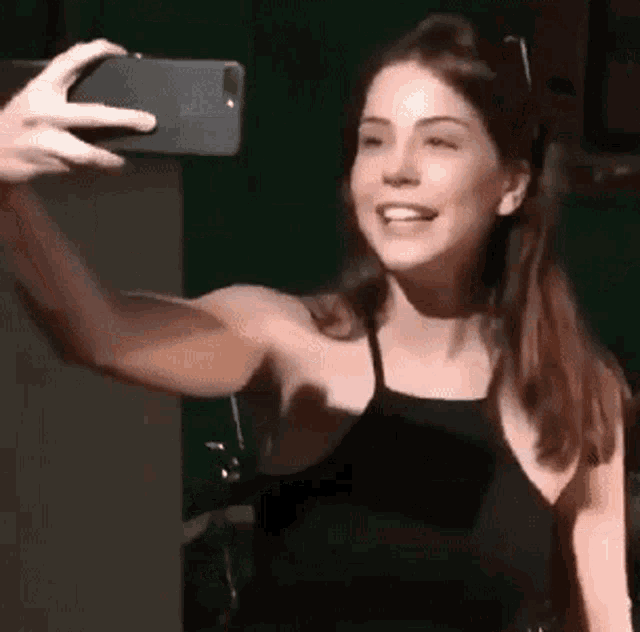 a woman in a black tank top is taking a selfie with a cell phone .