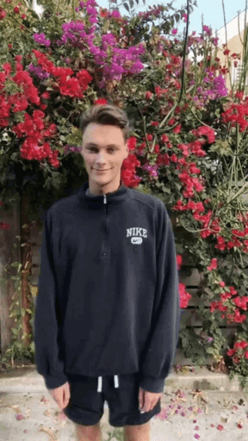 a young man wearing a black nike sweatshirt stands in front of a bush of flowers