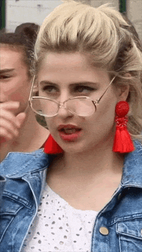 a blonde woman wearing glasses and red tassel earrings looks at the camera
