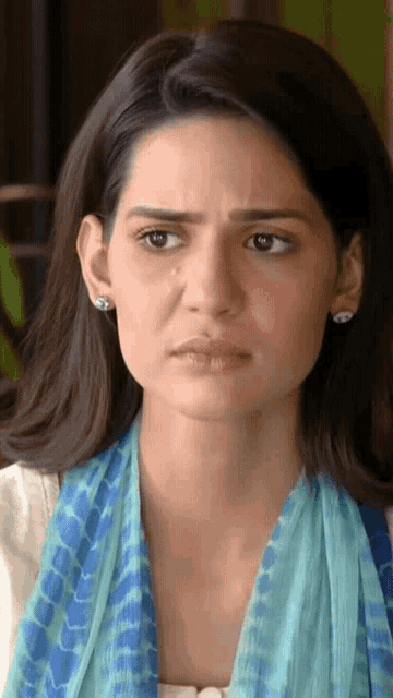 a woman wearing earrings and a blue scarf looks at the camera with a serious look on her face