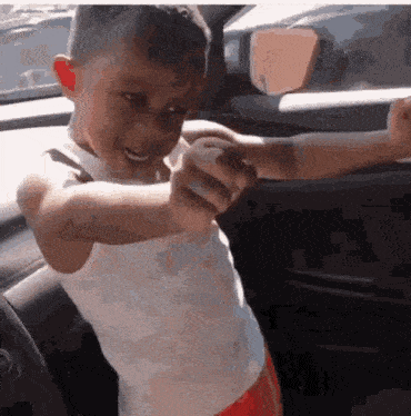 a young boy in a white tank top and red shorts is pointing at the camera .