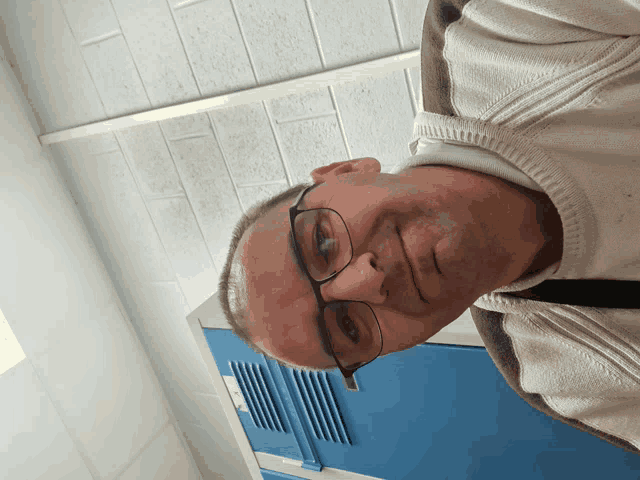 a man wearing glasses and a white sweater stands in front of blue lockers