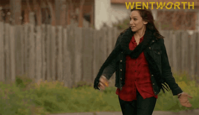 a woman in a red shirt and black jacket is walking in front of a wooden fence and the word wentworth is visible