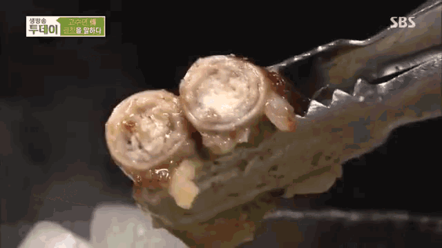 a close up of tongs holding a piece of food with sbs written on the bottom right