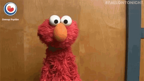 elmo from sesame street is standing in front of a wooden door and making a funny face .