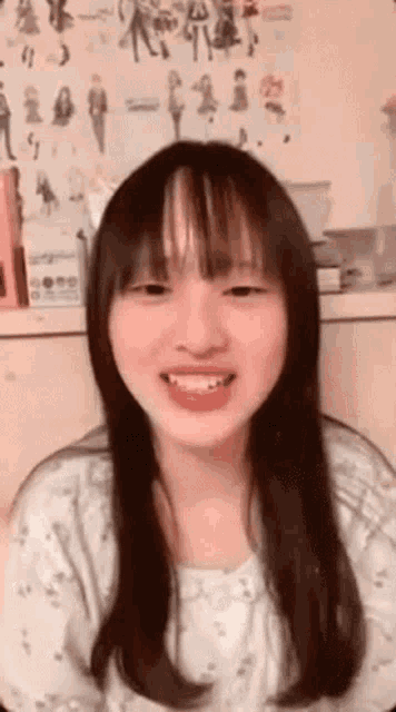 a young girl with long hair and bangs is smiling in front of a wall with drawings of people on it .