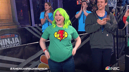 a woman in a green shirt with a lightning bolt on it is dancing in front of a banner that says american ninja warrior