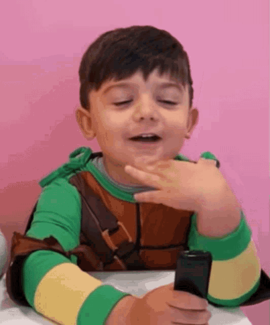 a young boy in a teenage mutant ninja turtle costume is holding a cell phone and smiling .