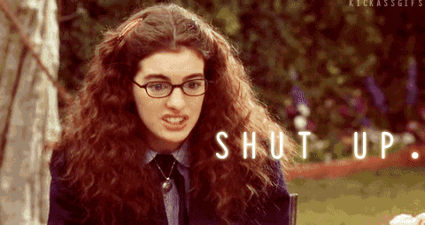 a woman with curly hair and glasses is sitting at a table with the words shut up written on the screen behind her .