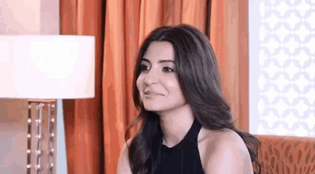 a woman in a black dress is sitting on a couch in front of a lamp and smiling .