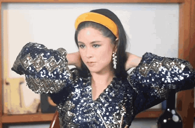 a woman wearing a blue sequined top and a yellow headband adjusts her hair