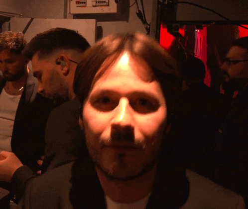 a man with a beard looks at the camera in a dark room with a few men