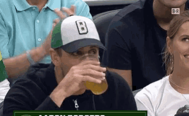 a man wearing a hat with the letter b on it drinks from a glass