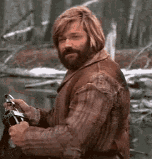 a man with a beard wearing a brown jacket is holding a fishing rod in the woods .