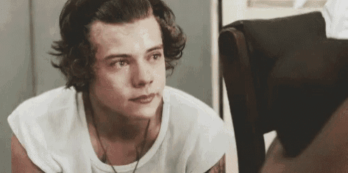 harry styles is wearing a white shirt and sitting in a chair looking at something .