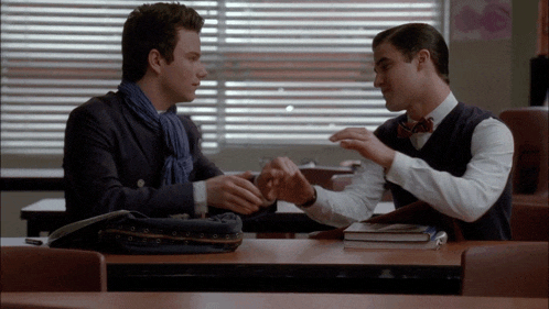 two men are sitting at a table holding hands and talking