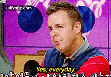 a man in a red and blue shirt is sitting in front of a microphone and saying yes , everyday .