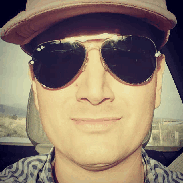 a man wearing a hat and ray-ban sunglasses smiles