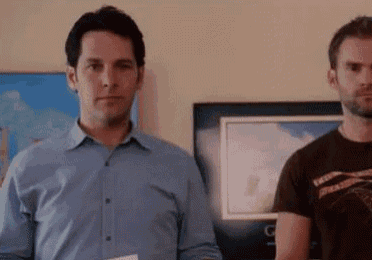 two men are standing next to each other in a room . one of the men is holding a piece of paper .