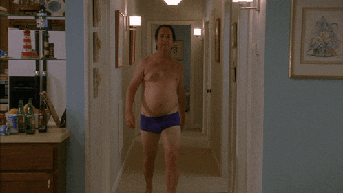 a shirtless man wearing purple swim trunks stands in a hallway