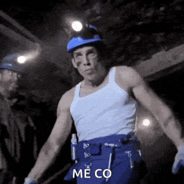 a man wearing a hard hat and a white tank top is standing in a dark room and says me co .